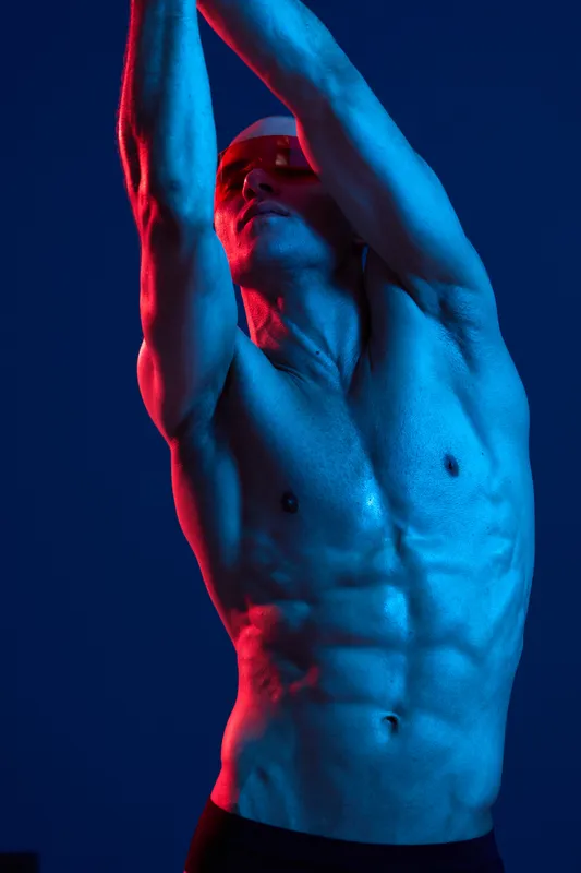 Athletic man striking a pose with dramatic lighting, showcasing toned physique and strong expression, set against a vibrant blue background.