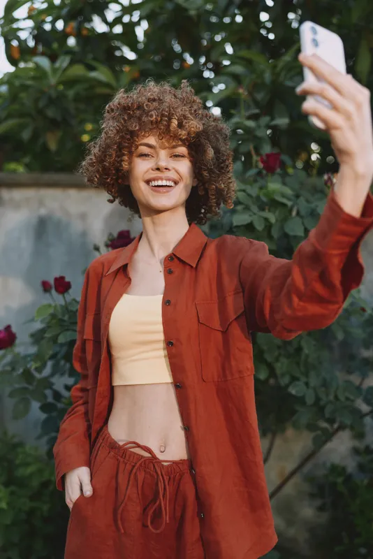 Young woman with curly hair takes a sunny selfie outdoors, wearing a red shirt over a beige crop top, exuding casual confidence and joy