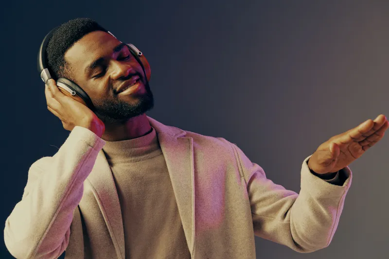 Joyful young African man enjoying music with headphones, wearing a stylish outfit, against a colorful gradient background emphasizing positive emotions and leisure