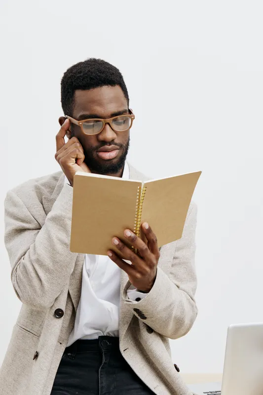 Thoughtful young African American man wearing glasses, beige blazer and white shirt, holding a notebook and reading carefully, desk with a laptop in front, neutral background, indoor office setting. B