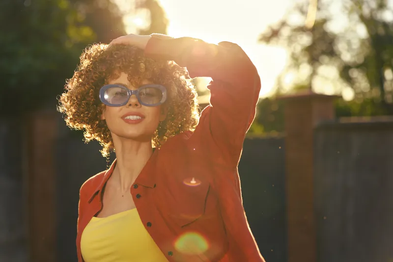 A stylish woman outdoors in bright sunlight wears a yellow top and red jacket, posing with sunglasses. The scene conveys carefree summer style and casual fashion in a sunlit setting