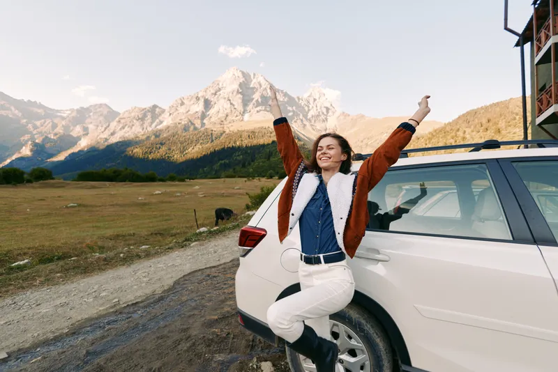 Woman, car, mountains, travel, nature, road, happy, outdoors. Female traveler leans on white SUV, stretching arms and smiling in a mountain meadow during a scenic countryside road trip at golden hour.