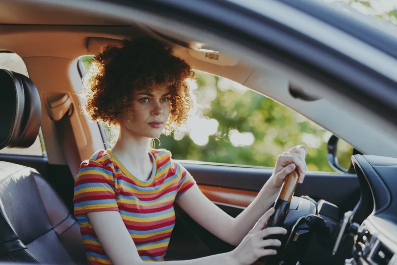Woman driving a car with striped shirt sits in the interior, daylight streaming through windows, capturing a portrait style lifestyle moment with retro film color and calm urban commute vibe