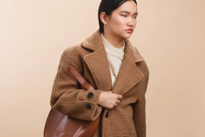 Asian woman wearing a warm brown wool coat stands against a soft beige backdrop, exuding calm confidence and professional style. Her composed expression conveys focus and poised presence.