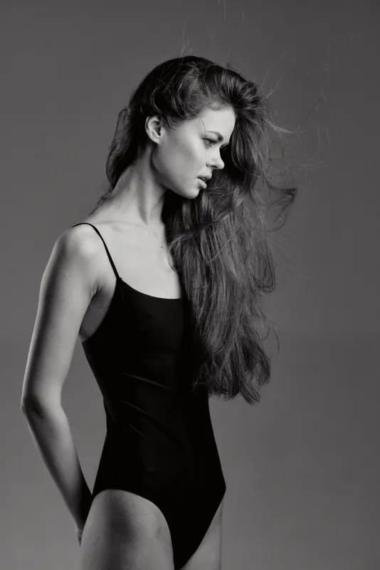 Black and white portrait of a woman with long hair wearing a black bodysuit looking down in studio lighting.