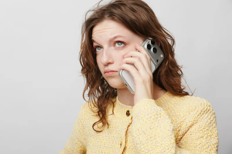 Young woman in yellow sweater looks worried while talking on smartphone against plain background. Concept of stress, communication, and emotional concern in everyday life.
