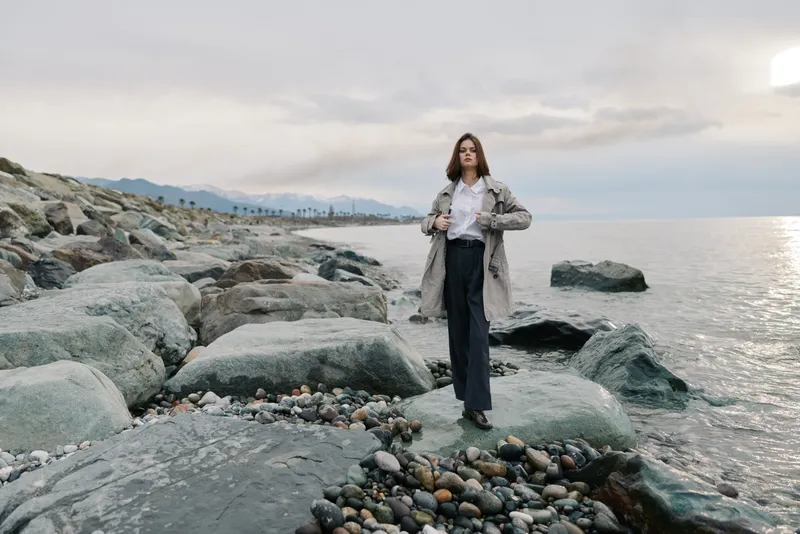 Young woman in coat stands on rocky shore near calm sea during cloudy day. Concept for fashion, nature, outdoor, and serene lifestyle with casual stylish outfit and natural background.