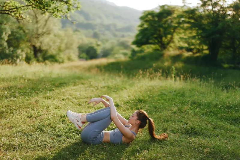 Fit young woman doing abdominal exercises in a sunny green park, showcasing strength and wellness with an outdoor backdrop