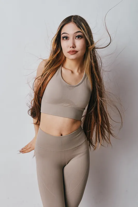 Young woman with long hair posing in beige fitness outfit with high waist leggings and crop top isolated on white background for active lifestyle and sportswear fashion.