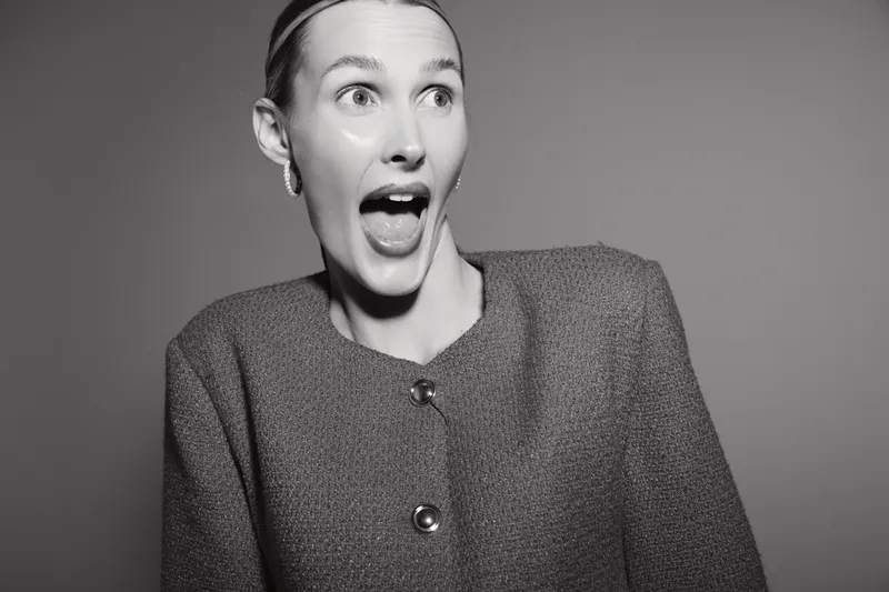Surprised white woman with light skin and short hair in textured jacket looks excited against plain background.