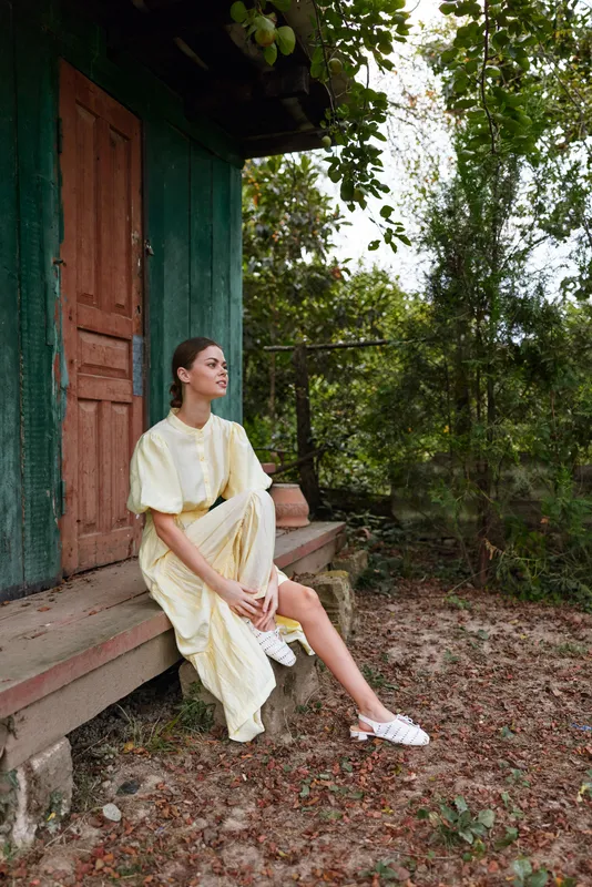 Woman on porch in a flowing dress sitting by a rustic countryside house in nature, relaxed pose with authenticity inclusivity, quiet moment, casual style and contemplative mood.