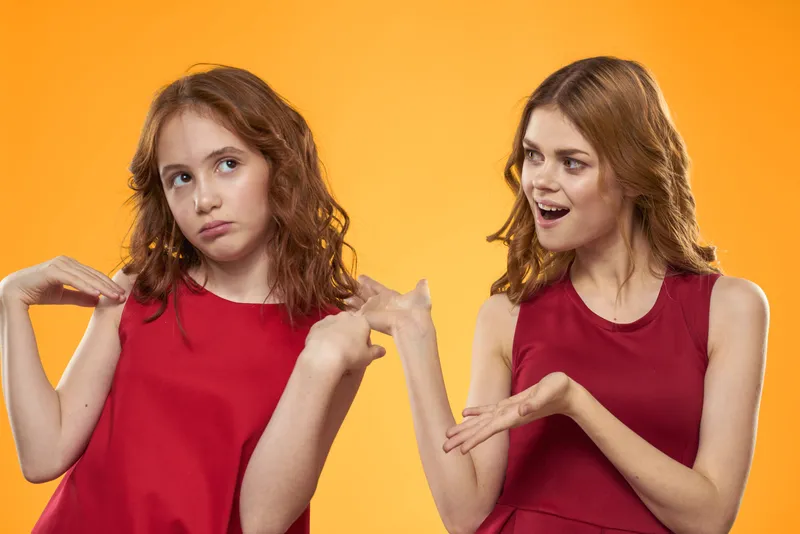 Две рыжеволосые девушки в красных платьях жестикулируют на оранжевом фоне