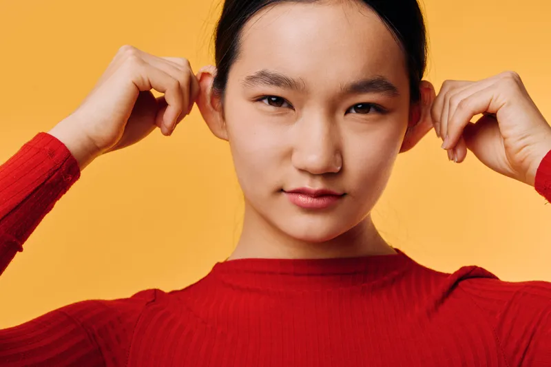 Portrait of young woman in red sweater against orange background adjusting hair with hands, looking directly at camera with soft natural expression in studio lighting.