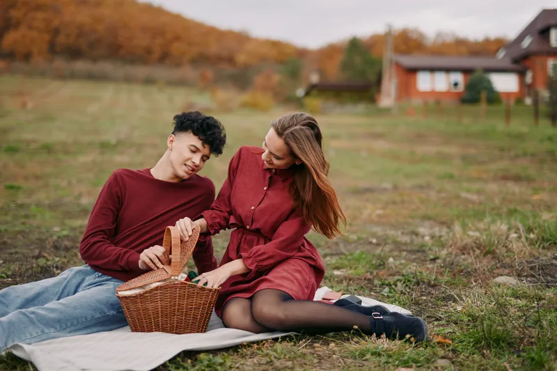 Two people sit on a blanket in an open field sharing a basket, a moment that feels authentic and credible, warmth in simple gesture and natural smiles amid autumn light.