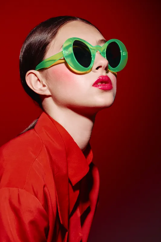 Fashionable young woman wearing bright green sunglasses poses against vibrant red background. Bold makeup with pink blush and intense red lipstick completes beauty creative style.