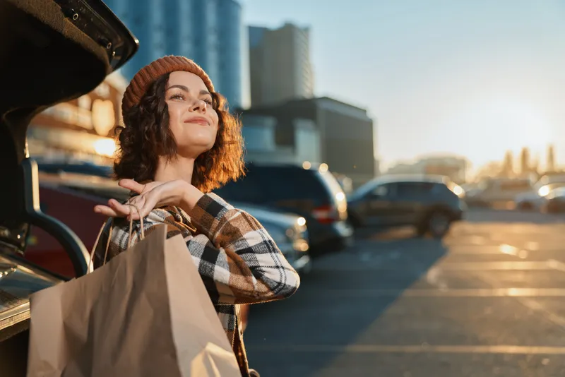 woman shopping car parking bags golden candid lifestyle authenticity mindful living emotional storytelling in urban sunset scene, cozy beanie portrait with smile and trunk full of retail purchases