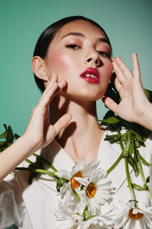 Stylish woman with red lipstick and radiant skin posing with white flowers on a green pastel background. Creative beauty and fashion concept with vibrant makeup.