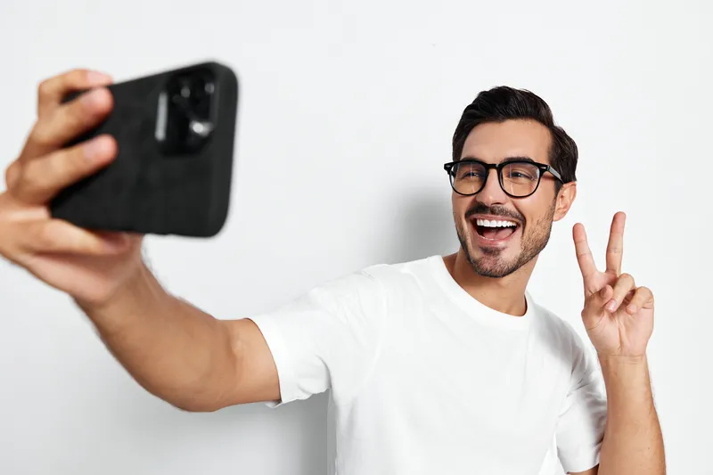 Smiling man with glasses taking selfie with mobile phone, showing peace sign, isolated on plain light background. Fun and lifestyle concept.