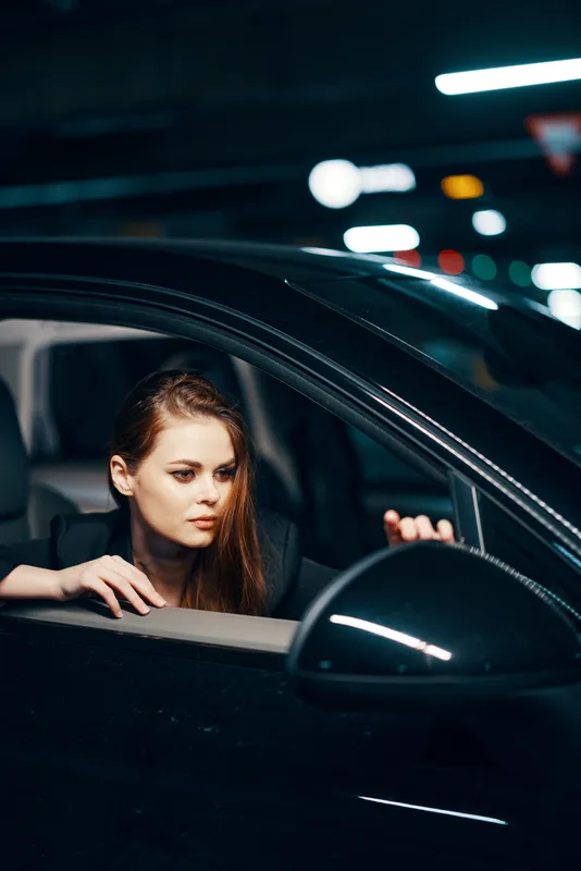 Вертикальная фотография, снятая сбоку ночью: женщина сидит в чёрном автомобиле, смотрит в окно и тянется к боковому зеркалу, чтобы поправить его
