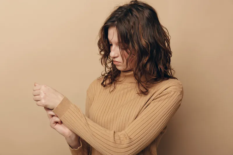 Young woman with curly hair wearing brown sweater looks down at her wrist with concern. Neutral beige background. Concept of discomfort, pain, or skin irritation.