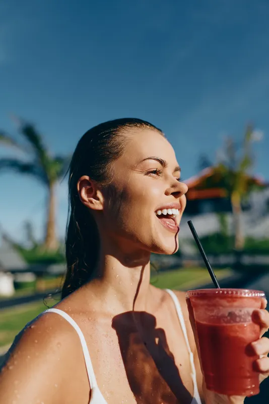Smiling woman with a refreshing drink outdoors under bright sunshine, close-up of a red smoothie in a glass with a straw, palm trees in the background, lifestyle moment of health, hydration and
