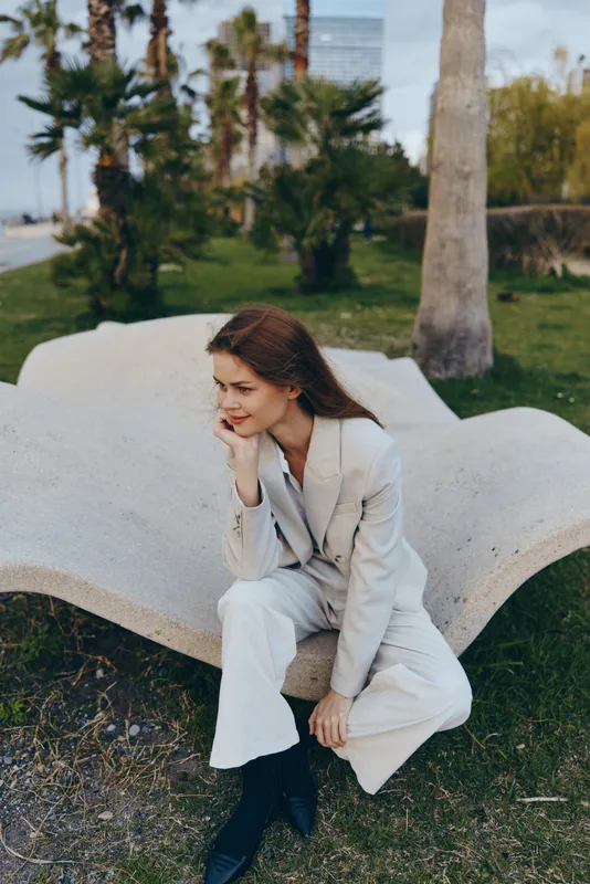 woman in light beige business suit thoughtfully sitting outdoors near palm trees autumn season urban environment