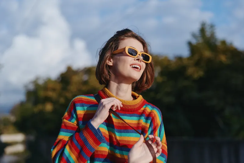 Woman smiling in a colorful striped sweater and yellow sunglasses, young woman enjoying outdoor portrait in park, short bob hair, casual outfit, looking up, joyful travel lifestyle and fashion moment.