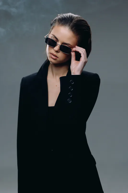Stylish woman wearing sunglasses and black blazer with serious expression and dark background lighting.