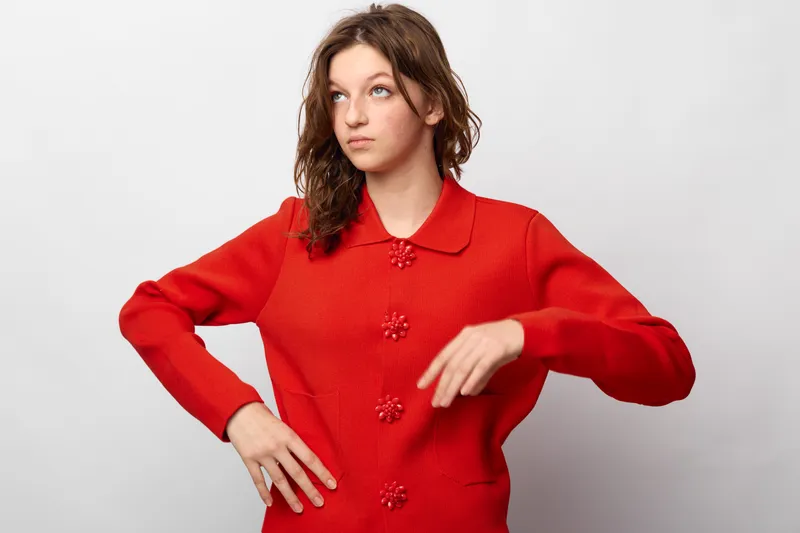 Young woman in red sweater with decorative buttons poses with hand on hip and looks away. Studio portrait on white background. Concept of fashion, casual style, and confidence.