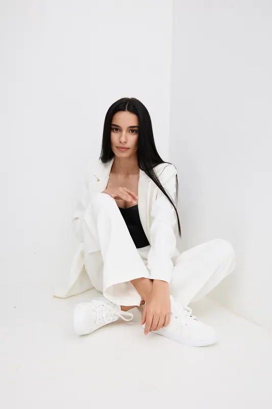 young woman sitting on white floor wearing white suit and sneakers, long dark hair, casual style, minimalistic bright background in studio setting