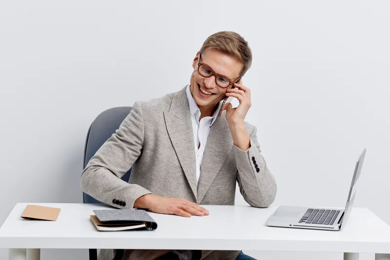 Professional smiling man in beige suit jacket working at modern office desk with laptop, notebook, and smartphone, looking happy and confident, isolated on plain white background, studio interior, bus