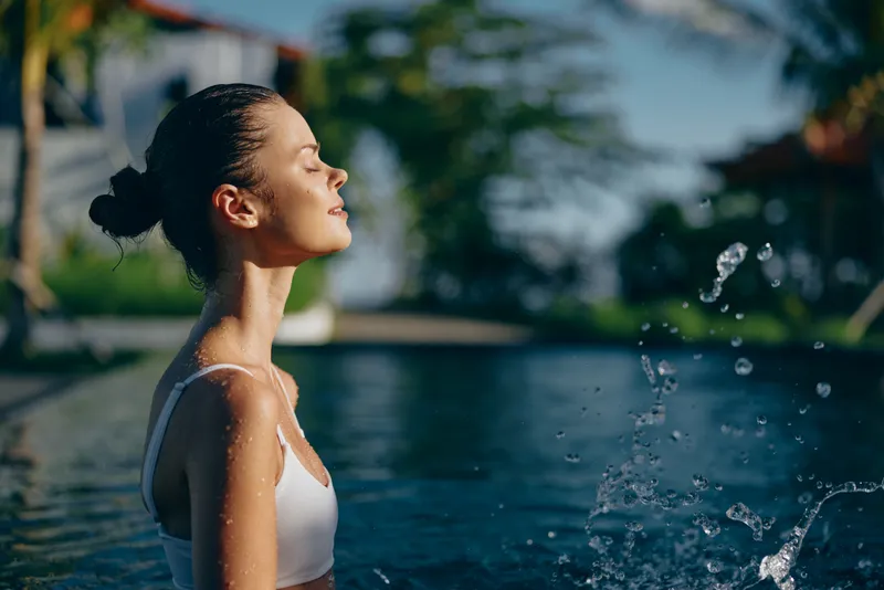 A woman at a sunlit poolside, eyes closed, enjoying a breezy outdoor moment as water sprays gently, conveying relaxed lifestyle and summer calm amid lush greenery and bright natural colors