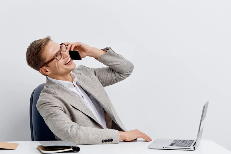 Professional woman in business suit talking on mobile phone at office desk with laptop, glasses, and notebook, smiling and cheerful, working in modern corporate environment, lifestyle concept, indoors