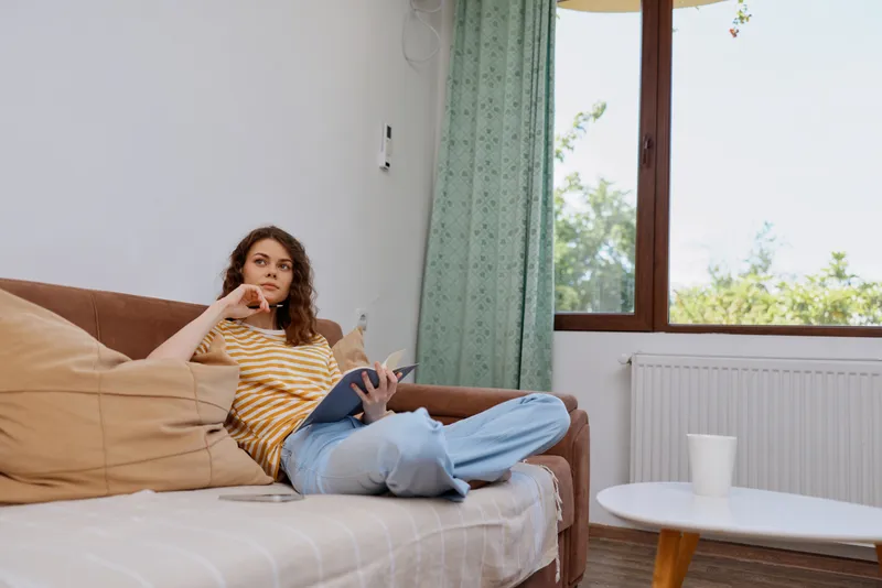 Casual woman relaxes at home on a sofa, reading tablet and resting, wearing striped shirt and jeans, cozy living room with natural light, thoughtful moment, and a window view adding warmth to the