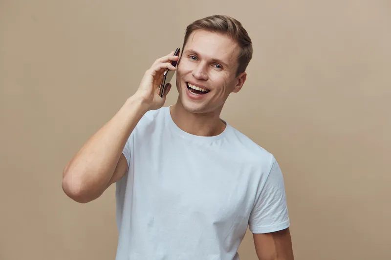 Smiling man talking on mobile phone isolated on beige background. Casual young male enjoying conversation, cheerful expression, friendly vibe, studio shot, portrait, lifestyle scene, relaxed style.