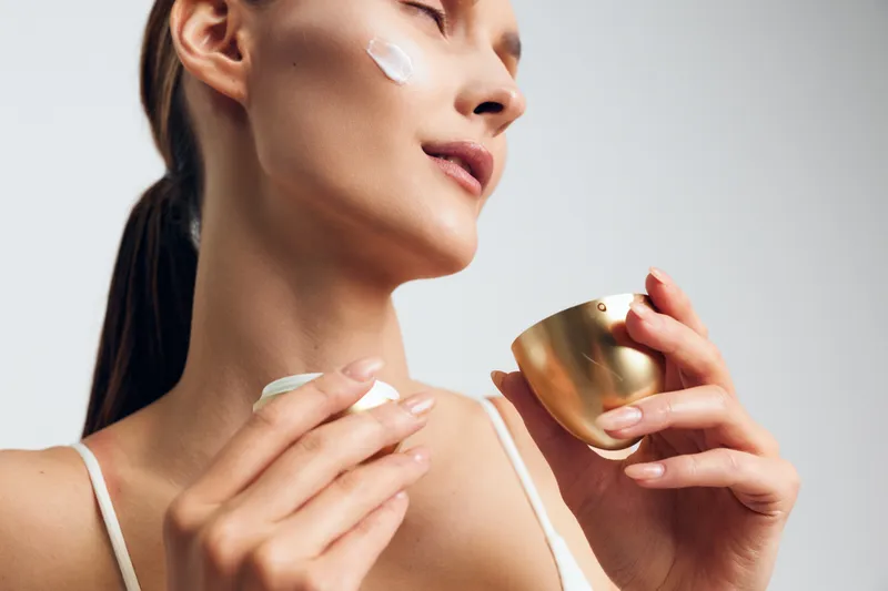 Young woman applying face cream with closed eyes, smooth skin, relaxed expression, soft lighting, beauty routine, skincare product in golden jar, minimalistic background