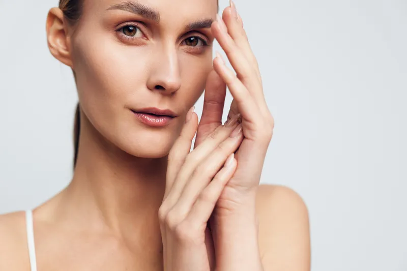 A close-up portrait of a young woman with clear skin gently touching her face with both hands against a plain light background. Concept for skincare, beauty, and natural look.