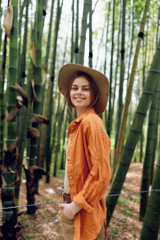 Woman portrait in a bamboo forest, smiling and wearing a wide hat and orange shirt. Natural outdoor scene with green foliage, relaxed pose, sunlight, trail and serene lifestyle feeling.