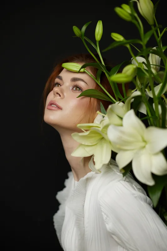 Elegant woman with red hair and white dress, surrounded by lilies, conveying beauty and grace in a dark background, perfect for floral and fashion concepts
