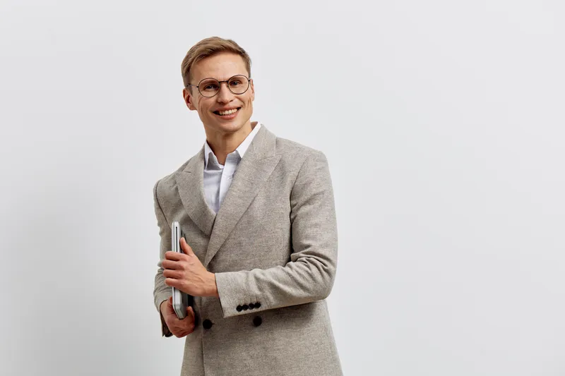 Professional young smiling man in smart casual clothing holding notebooks and a pen, looking confidently at camera against plain light gray background, studio portrait. Business lifestyle concept