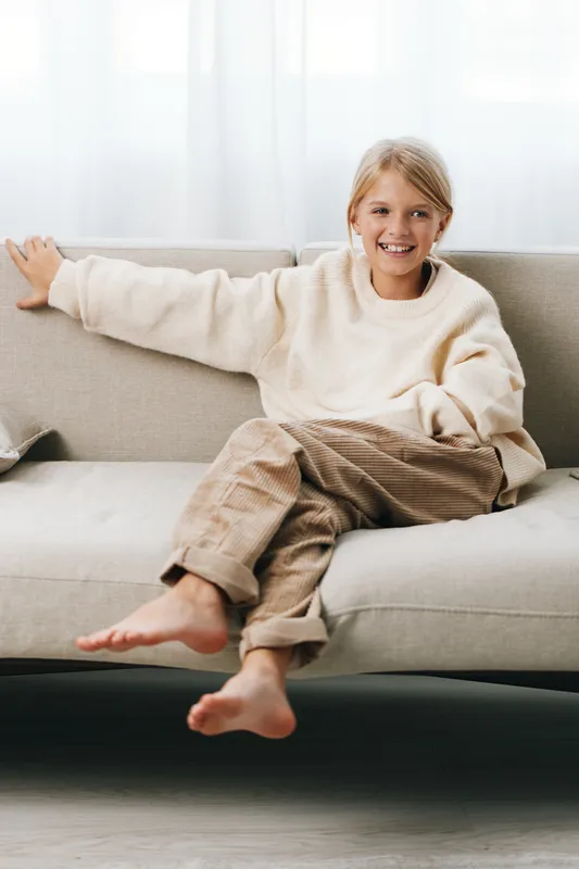 Smiling child sits comfortably on a light sofa, wearing a cream sweater and beige pants, feet bare, relaxed pose conveying warmth, comfort, and casual home style.