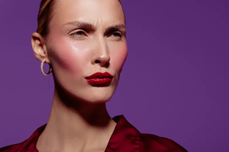 Close-up of a woman with glossy red lips and pink blush on cheeks wearing gold hoop earrings and a red blouse. Purple background highlights her confident and stylish look.