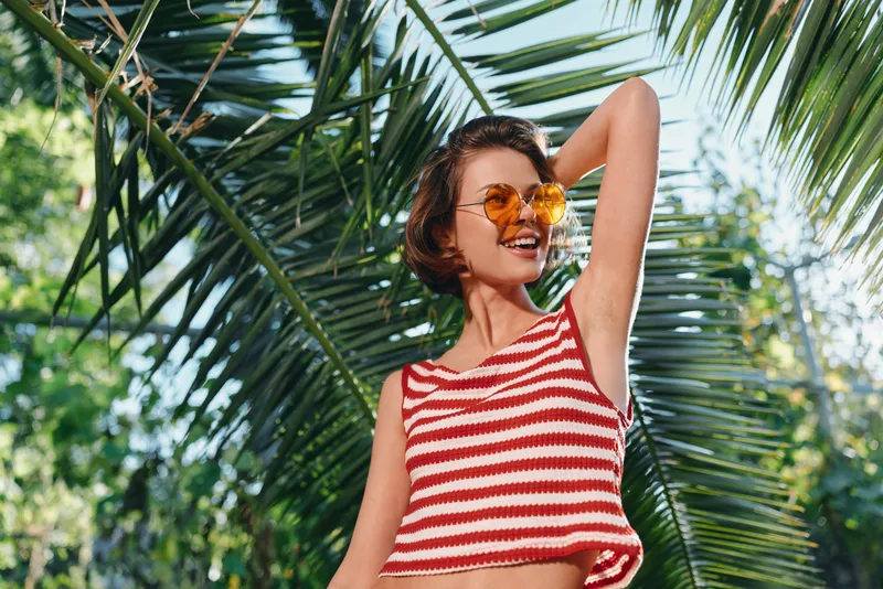 A cheerful woman wearing a striped crop top and sunglasses stands in a sunny tropical garden, posing with confidence and a carefree smile under lush palm leaves.