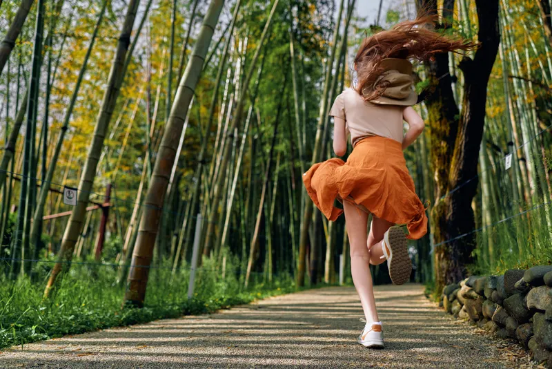 Woman running on a bamboo path in a green forest, motion captured from back view, orange skirt flowing and white sneakers on a sunlit trail among tall stalks.