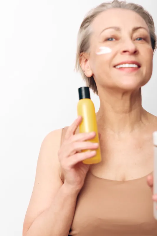 Smiling mature woman applying face cream with yellow bottle in hand, showing skincare routine and wellness. Bright clean background highlights healthy skin and self-care concept.