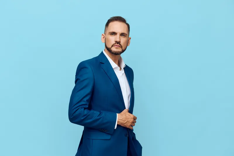 A focused male business professional stands against a solid blue background, exuding confidence and determination as he adjusts his suit, showcasing readiness, resolve, calm ambition, and executive