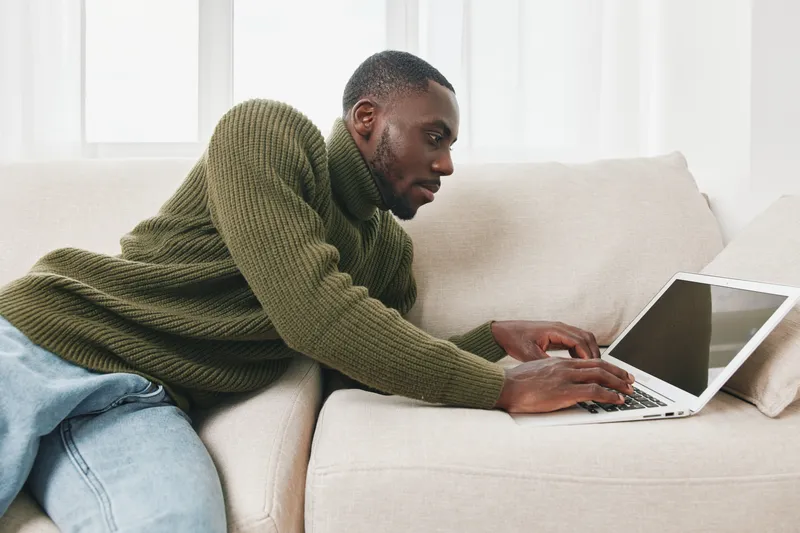 Young Black man in cozy green sweater working on laptop at home, showcasing focus and creativity in a modern, bright living room setting