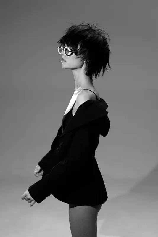 Black and white portrait of a stylish woman with short hair wearing sunglasses and an off-shoulder jacket.