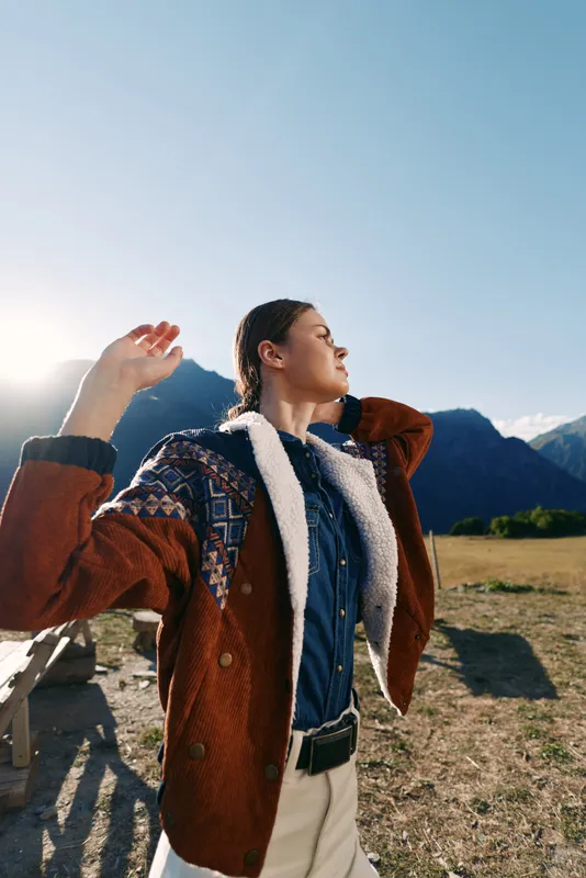 Woman portrait in mountains outdoors wearing a warm patterned jacket, basking in golden sunlight in an open field. Fashionable traveler enjoying nature, fresh air and mountain landscape.
