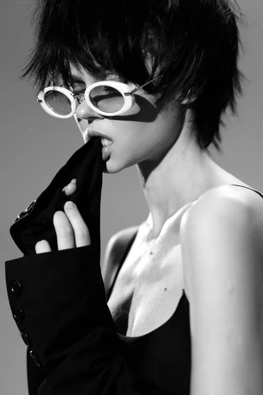 Black and white portrait of a stylish woman wearing white sunglasses and biting her black sleeve, expressing edgy and confident attitude.
